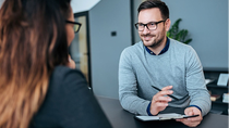 Zwei Personen sitzen an einem Tisch in einem modernen Büro und führen ein Gespräch, während sich die eine Person Notizen macht. Zwei Personen sitzen an einem Tisch in einem modernen Büro und führen ein Gespräch, während sich die eine Person Notizen macht.