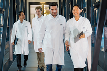 Vier Personen in weißen Laborkitteln gehen durch einen modernen Glasgang und symbolisieren Teamarbeit und Forschung in einem innovativen Umfeld. Vier Personen in weißen Laborkitteln gehen durch einen modernen Glasgang und symbolisieren Teamarbeit und Forschung in einem innovativen Umfeld.